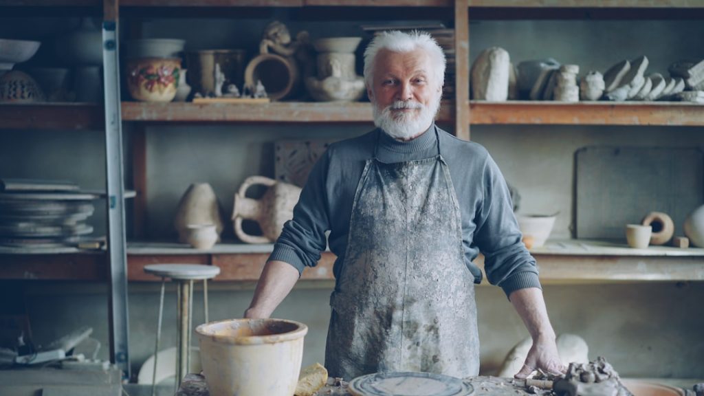 Portrait of senior silver-haired potter in muddy apron standing alone at table in workshop and looking at camera. Shelves with handmade vases and pots in background.