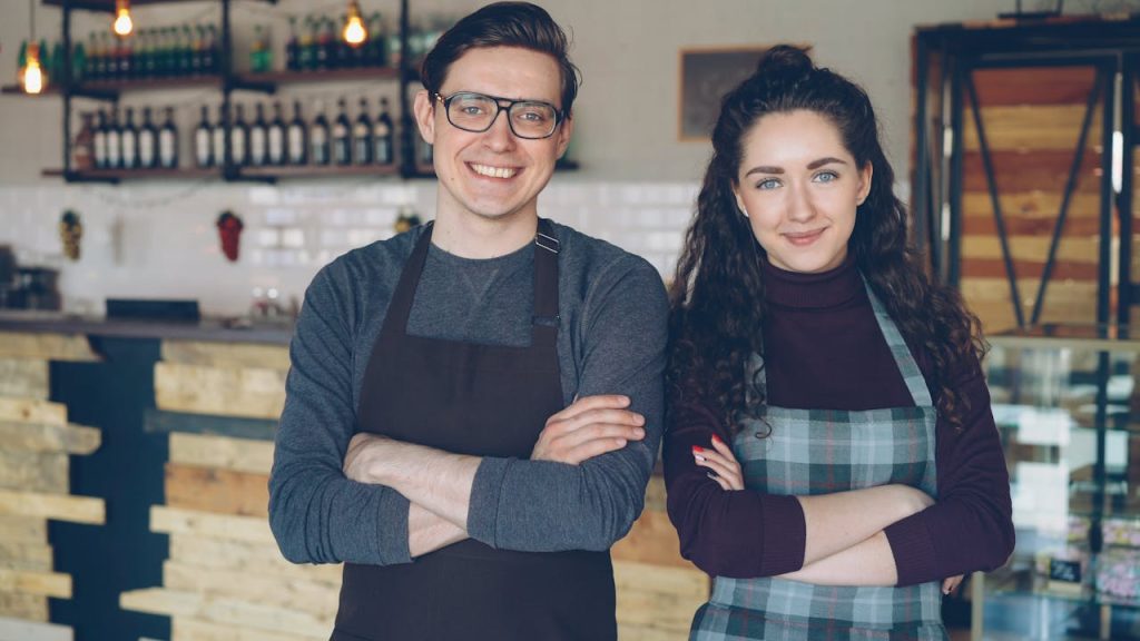 Two young cafe owners stand proudly in their modern cafe, smiling and wearing aprons.
